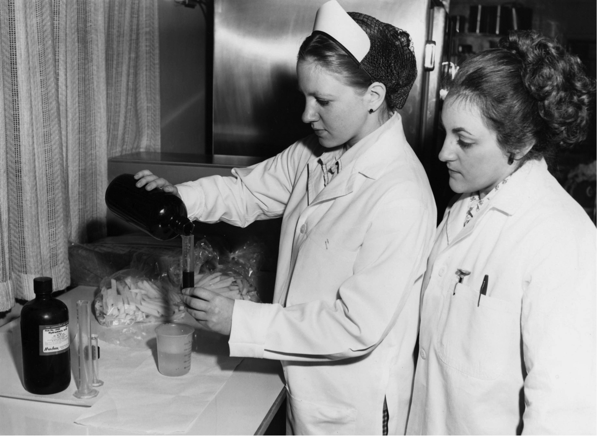 Two girls wearing lab coats measure a liquid in the cooking lab. One of them is wearing a hair net (1960.)