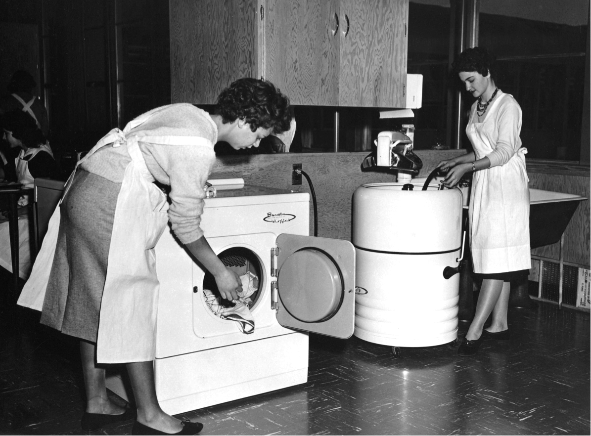 Two girls do laundry in home economics class. One student operates a wringer-type washing machine, the other, a dryer (1960.)
