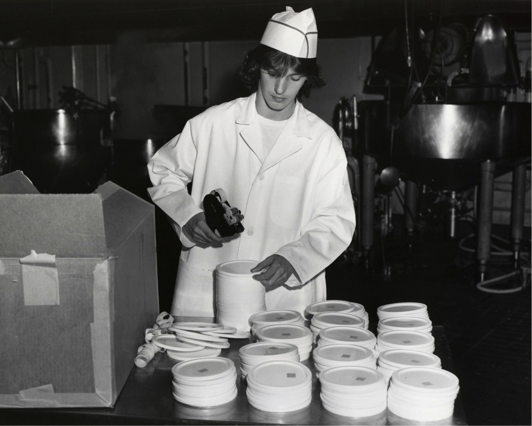 A ‘work experience’ student uses a labelling machine to label food containers at a White Spot restaurant (1974.)